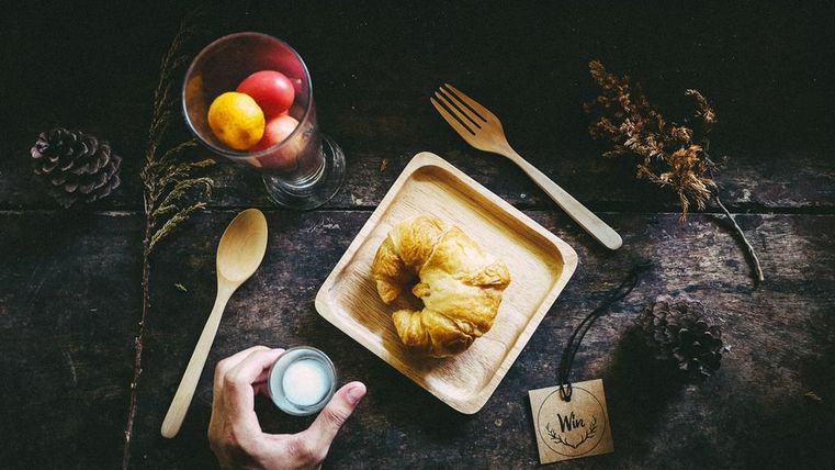 Une table rustique avec un plateau en bois sur lequel se trouvent une pâtisserie et une coupe en verre avec des fruits frais. Une main tient un petit verre tandis qu'une cuillère est posée à côté.