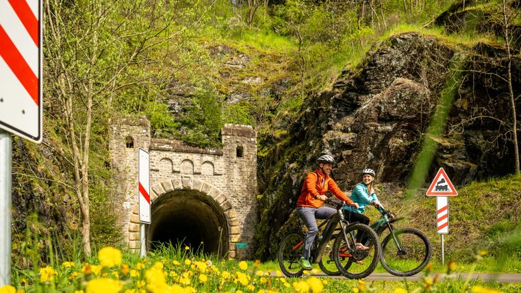 Radfahrer genießen die Natur vor einem alten Tunnel. Umgeben von grünen Wiesen und blühenden Löwenzahn.
