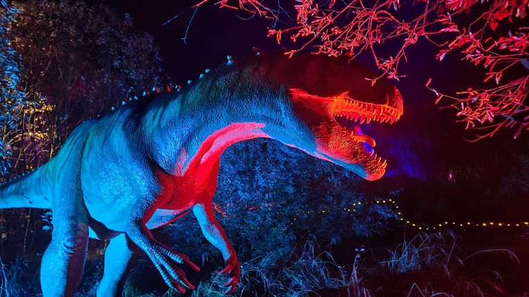 A glowing dinosaur stands in a dark environment, surrounded by colorful lights. The colors are intense and give the scene an exciting atmosphere.