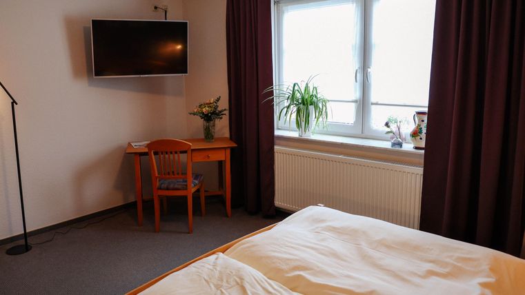 Ein einfaches Zimmer mit einem Bett, einem Tisch und einem Fernseher. Die Fenster lassen viel Tageslicht herein und es gibt eine Pflanze auf dem Tisch.