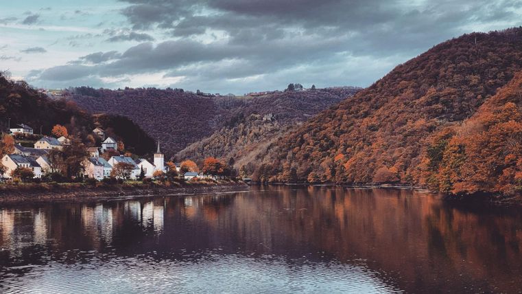 Eine ruhige Landschaft mit sanften Hügeln und herbstlichen Bäumen. Im Vordergrund spiegelt sich der Fluss in ruhigem Wasser.