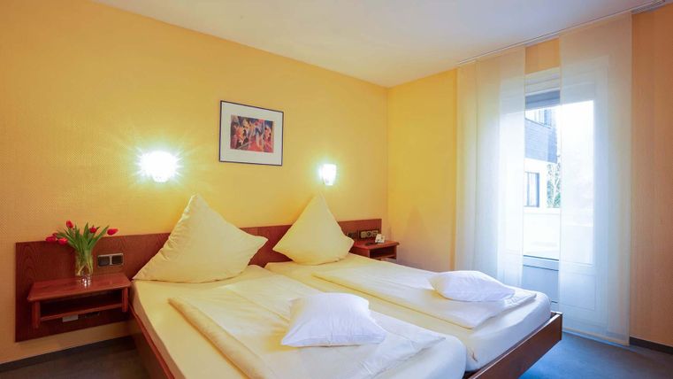 Ein helles Zimmer mit zwei Einzelbetten und gelben Wänden. Es gibt eine große Fensterfront und eine Blumendekoration auf dem Tisch.