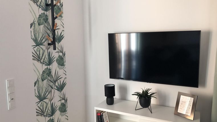 Een moderne woonkamer met een televisie aan de muur. Naast het tv-meubel staan een kleine lamp en een plant.