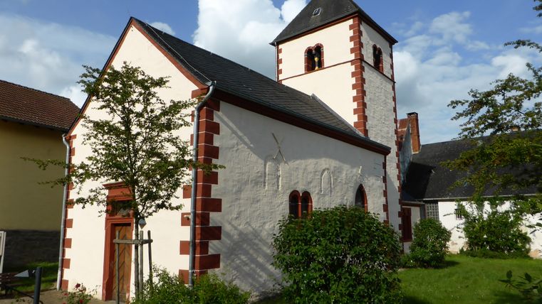 Kleine Kirche mit rotem Ziegelsteinmuster und Turm, umgeben von Bäumen und Büschen.