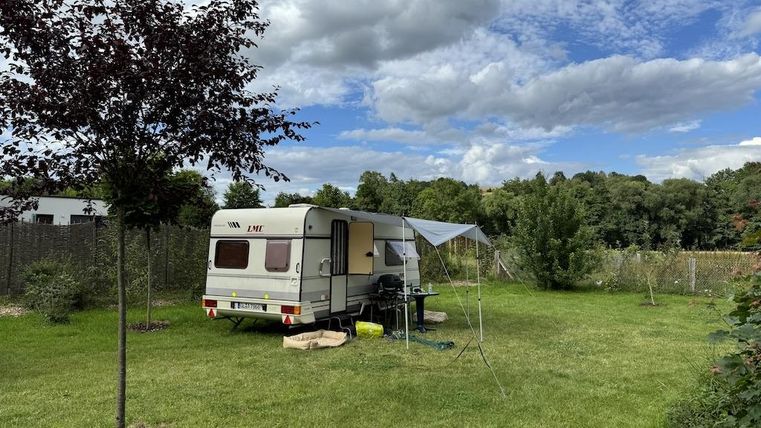 Een caravan staat op een groene weide, omringd door bomen en struiken. De lucht is gedeeltelijk bewolkt en de scène oogt vredig en uitnodigend.