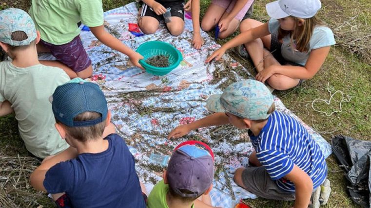 Eine Gruppe von Kindern sitzt auf einem Tuch und spielt mit verschiedenen Materialien. Sie verwenden eine Schüssel und wirken konzentriert und glücklich.