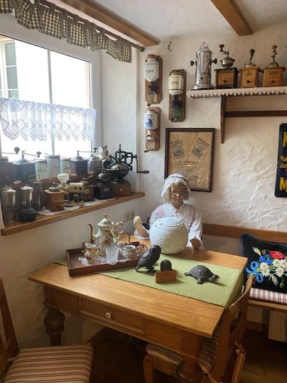 Ein gemütlicher Raum mit einem Tisch und verschiedenen dekorativen Gegenständen. Eine Puppe in traditioneller Kleidung sitzt in der Ecke und verleiht dem Raum einen nostalgischen Charme.