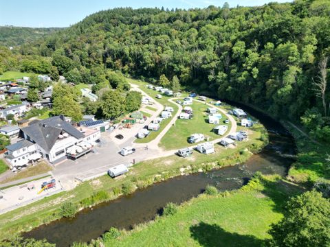 Ein Campingplatz neben einem Fluss, umgeben von Bäumen und Hügeln. Mehrere Wohnmobile und Zelte sind auf dem Gelände verteilt.