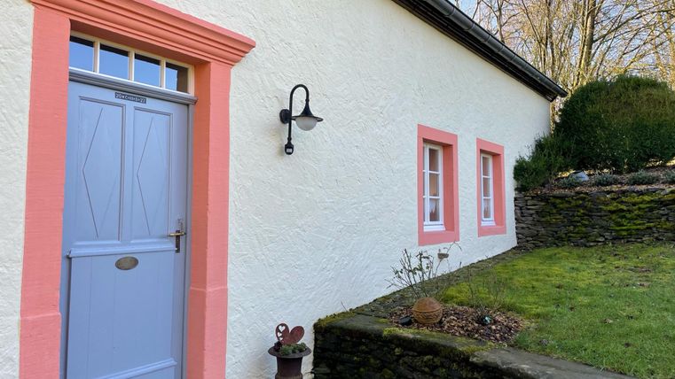 Ein gemütliches Haus mit einer hellen Fassade und einer blauen Tür. Vor dem Haus befindet sich ein kleiner Garten mit Steinen und grünen Pflanzen.