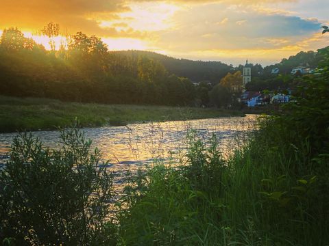 Ein ruhiger Fluss fließt durch eine grüne Landschaft. Im Hintergrund strahlt die Sonne hinter sanften Hügeln und Gebäuden.