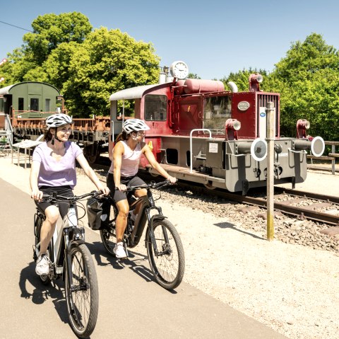 Zwei Radfahrerinnen auf einem Radweg neben einer alten Lokomotive im Eisenbahnmuseum Pronsfeld, umgeben von grünen Bäumen., © Eifel Tourismus GmbH, Dominik Ketz