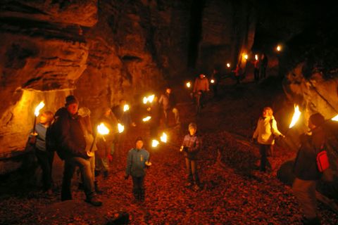 Eine Gruppe von Menschen geht mit Fackeln durch eine dunkle Höhle. Der Boden ist mit Laub bedeckt, und das Licht der Fackeln beleuchtet die Umgebung.