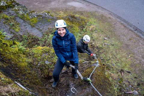 Zwei Personen klettern an einer steilen, bewachsenen Felswand. Beide tragen Helme und sind mit Kletterausrüstung ausgestattet.