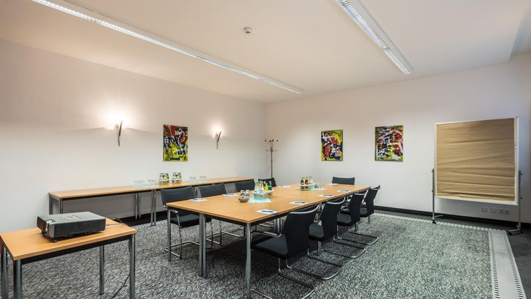 Ein moderner Besprechungsraum mit einem großen Tisch und mehreren Stühlen. An den Wänden hängen bunte Bilder, und es gibt Getränke auf dem Tisch.