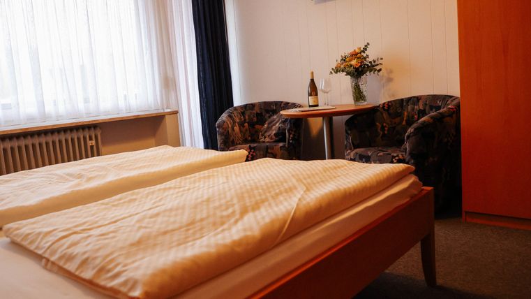 Ein gemütliches Schlafzimmer mit zwei Betten und weichen Decken. Im Hintergrund befinden sich zwei Sessel, ein Tisch und eine Vase mit Blumen.