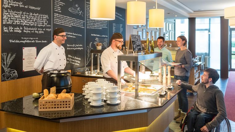 Eine moderne Kantine mit Köchen, die Essen anrichten. Gäste werden freundlich bedient, darunter auch eine Person im Rollstuhl.