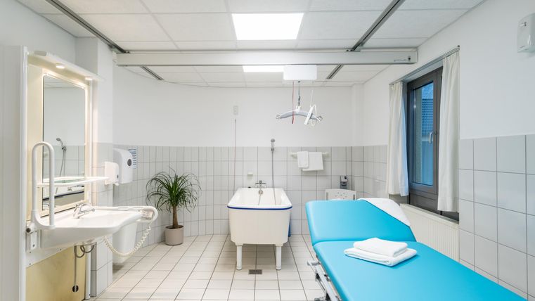 Ein modernes Behandlungszimmer mit einem blauen Untersuchungstisch und einer weißen Badewanne. Der Raum ist hell und freundlich mit Pflanze und großen Fenstern.
