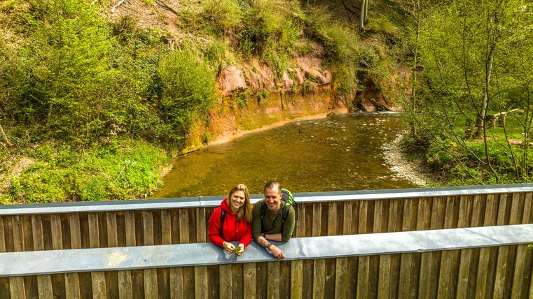 Ein Paar posiert an einem Holzgeländer in der Natur. Im Hintergrund fließt ein kleiner Fluss, umgeben von Bäumen und grünem Laub.