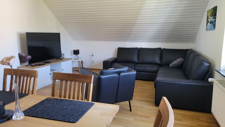 Ein modernes Wohnzimmer mit einem großen, schwarzen Sofa und einem Flachbildfernseher. Der Raum hat einen holzverkleideten Boden und eine helle, einladende Atmosphäre.