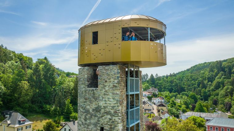 Ein moderner Aussichtsturm aus Stein mit einer goldenen Plattform. Umgeben von grünen Hügeln und kleinen Gebäuden.