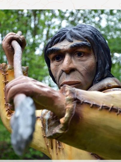 Nahaufnahme einer prähistorischen Figur mit Speer im Dinopark, umgeben von Bäumen.